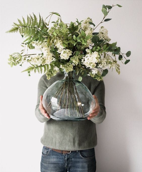 a person holding a vase with flowers in it and looking down at the ground while standing against a wall