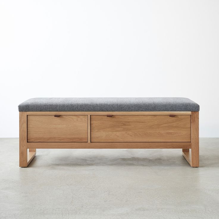 a wooden bench with two drawers and a gray cushion on the top, in front of a white wall