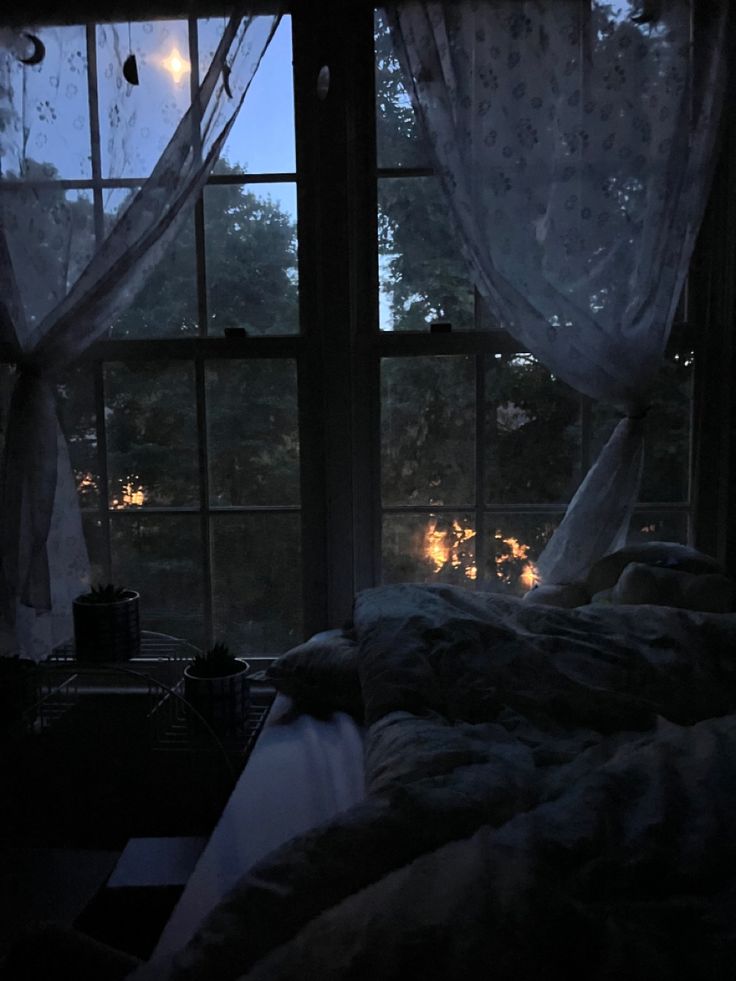 a bed sitting next to a window with curtains on it's sides and lights in the distance