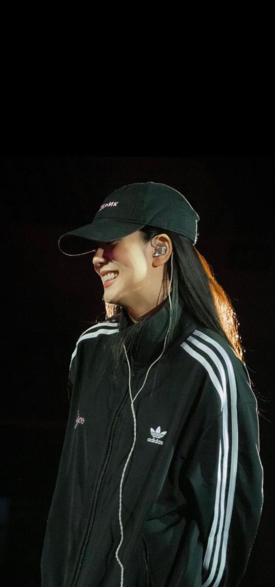 a woman with long hair wearing a black jacket and hat, standing in front of a dark background