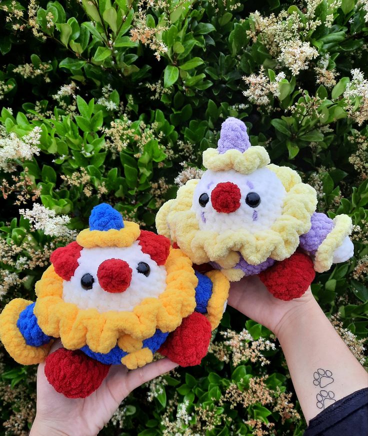 two crocheted clown faces are held in front of some bushes with white flowers