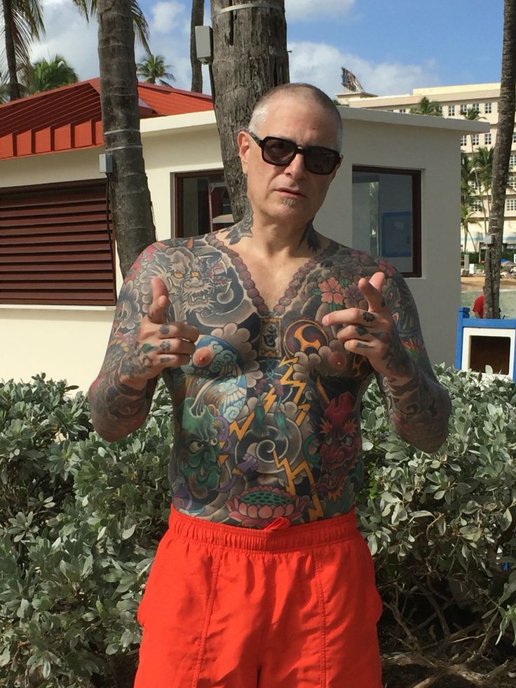 a man with tattoos standing in front of some trees and bushes, giving the peace sign