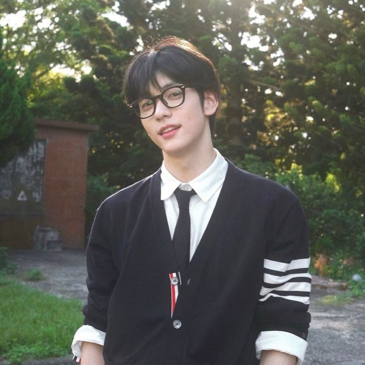 a young man wearing glasses and a black sweater is standing in front of some trees