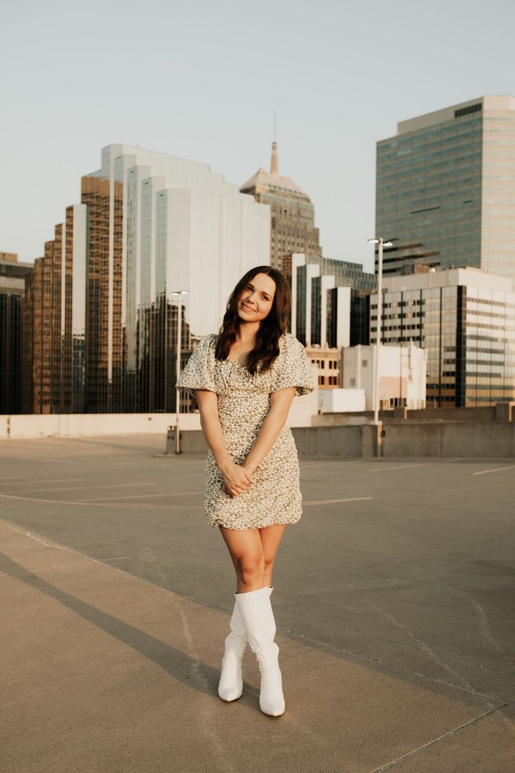 a woman standing in an empty parking lot wearing white boots and a dress with short sleeves