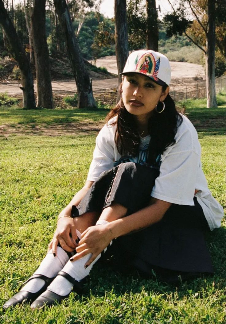 a woman sitting in the grass with her legs crossed wearing socks and a baseball cap