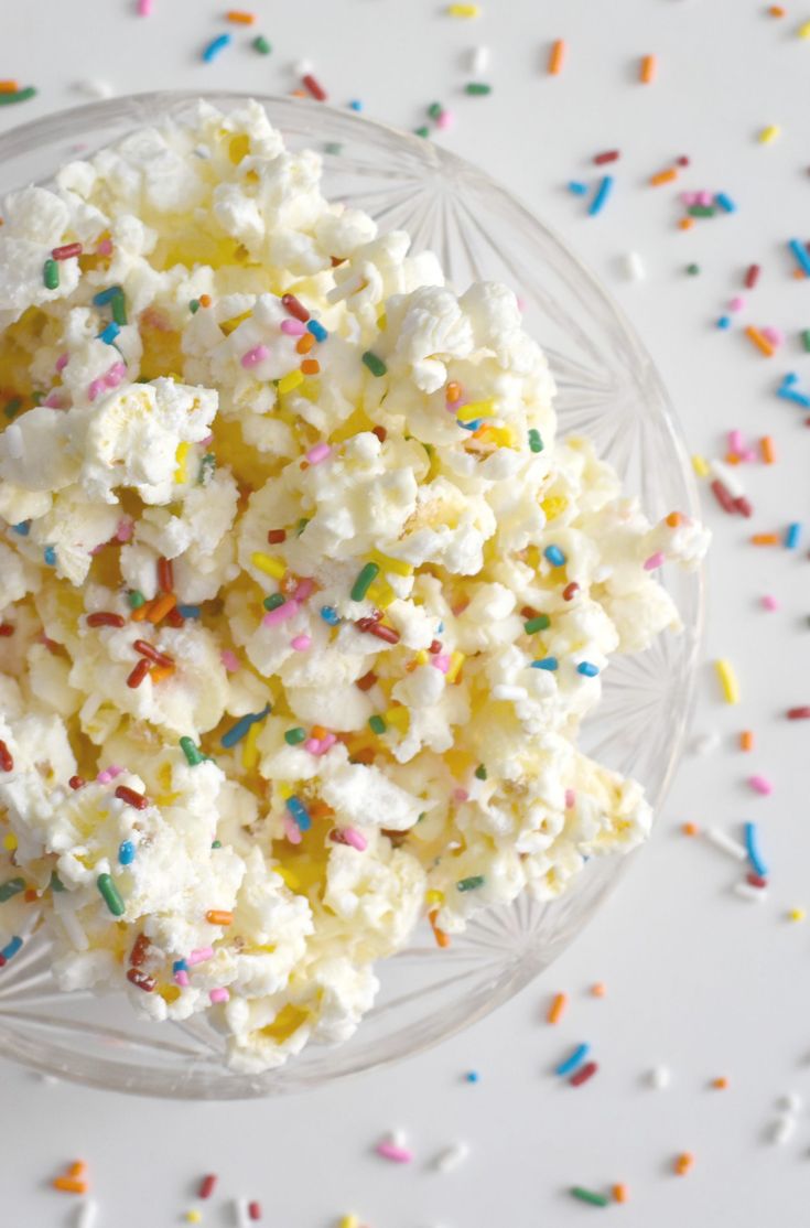 popcorn with sprinkles in a glass bowl on a white table surrounded by confetti and sprinkles