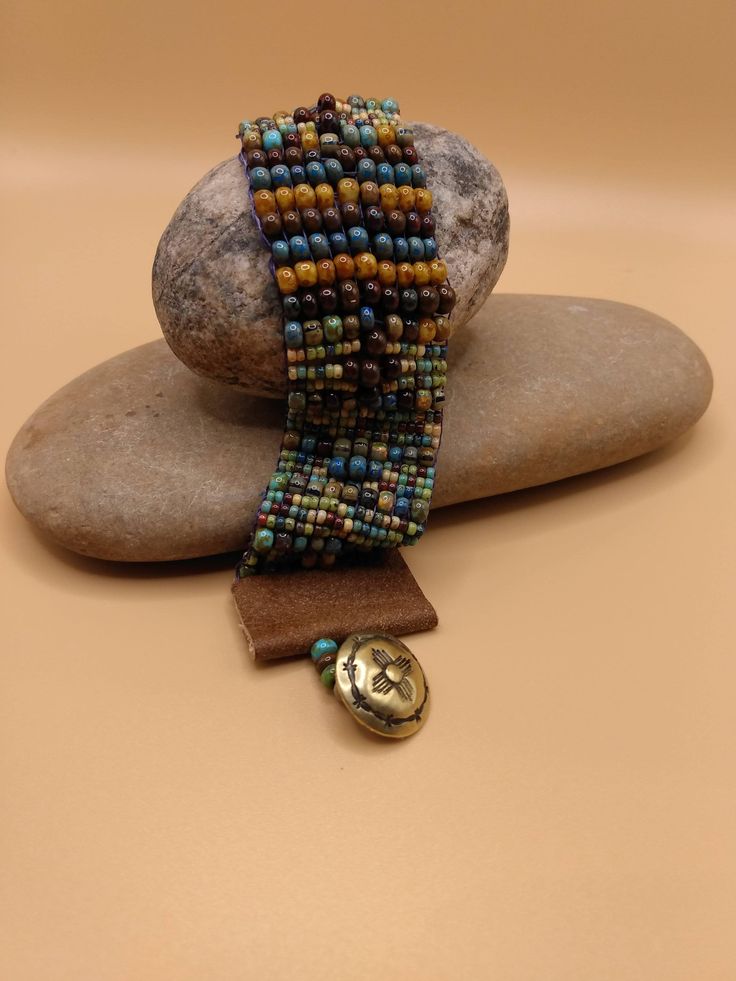 This beautiful One- of- a -Kind southwest style beaded bracelet is adorned with beautiful Picasso beads of varying colors of gold,  turquoise blue, smoky blue, brown, soft green, and burgundy.  The larger beads are 4 m. m.  Bohemian mix Opaque Picasso . The small ones are size 6 Aged Striped Czech Glass Rocaille seed beads.  The ends of the bracelet are finished with tan leather with a bronze southwest style button.  This bracelet is 7 inches long and 1 inch wide. This bracelet will fit 6-7 inch inch wrist .   I hope that you will love this very stylish bracelet!   ENJOY! Artisan Beaded Multicolor Wrap Bracelet, Artisan Multicolor Beaded Wrap Bracelet, Brown Wrap Bracelet With Colorful Beads For Festival, Earthy Turquoise Bracelets For Festival, Earthy Colorful Beaded Bracelets For Festivals, Earthy Style Colorful Beaded Bracelets For Festivals, Earthy Brown Beaded Wrap Bracelet, Bohemian Cuff Bracelet With Large Beads, Hippie Bracelet With Colorful Beads