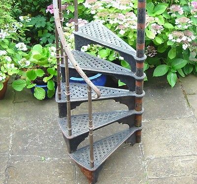 an old metal spiral staircase in front of some plants and flowers on the ground with potted plants behind it