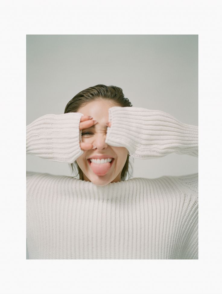 a woman covering her eyes and smiling with both hands behind her head, wearing a white sweater