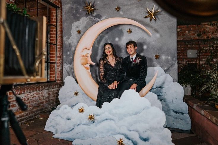 a man and woman sitting on top of a fake moon in front of a backdrop