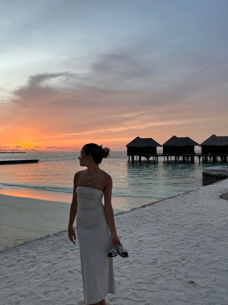 a woman in a white dress standing on the beach at sunset with some huts in the background