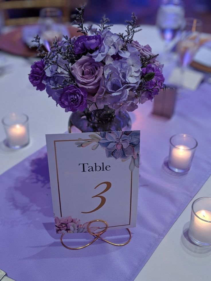 a table number with purple flowers in a vase and candles on the table at a wedding reception