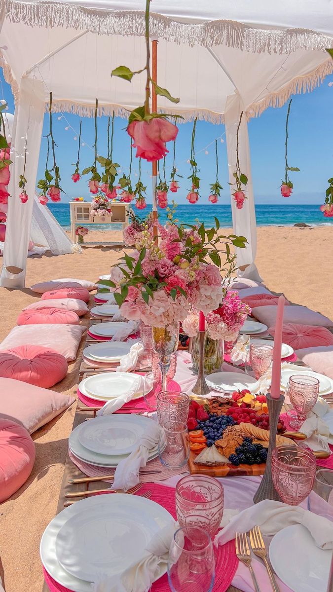 a table set up with plates and pink napkins for an outdoor dinner on the beach