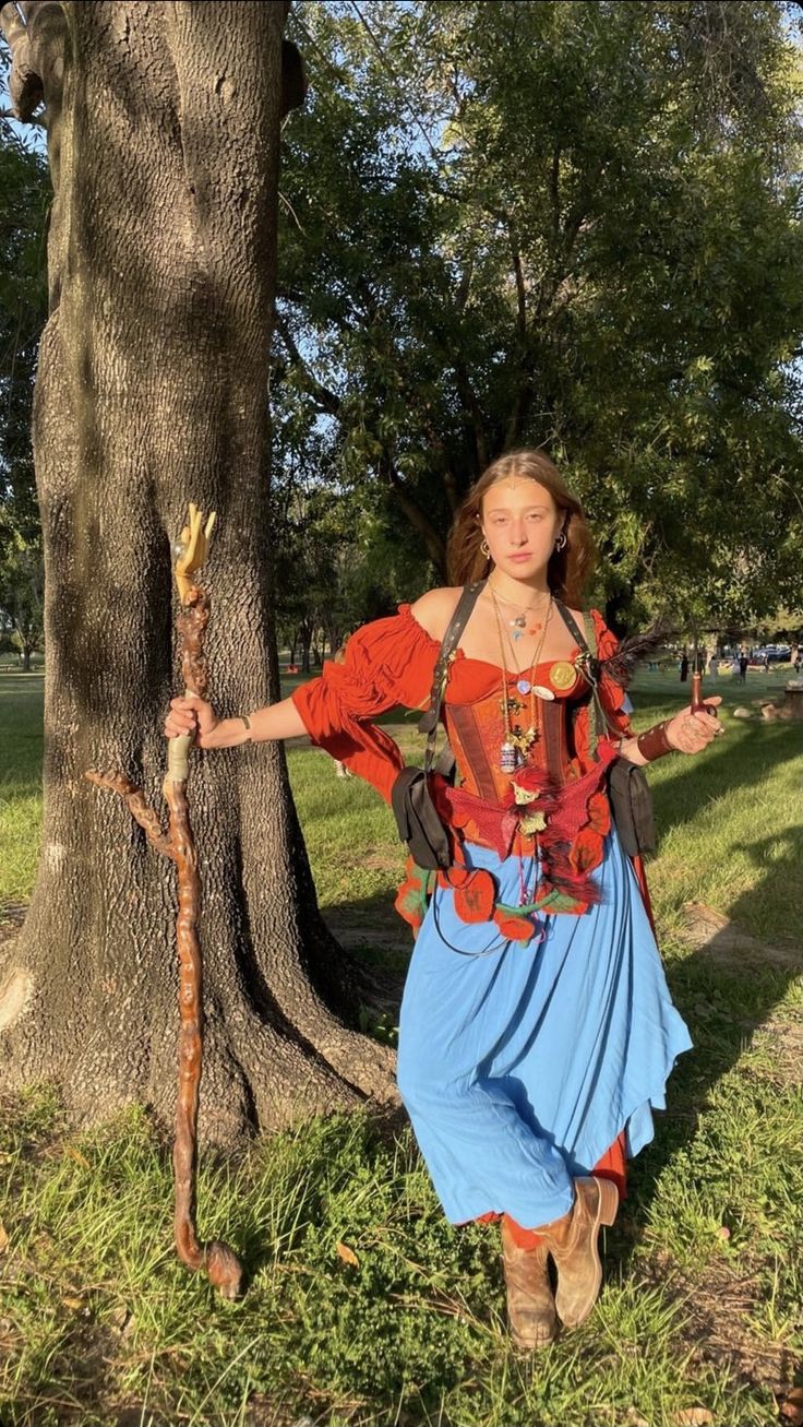 a woman standing next to a tree in a park wearing a costume and holding a stick