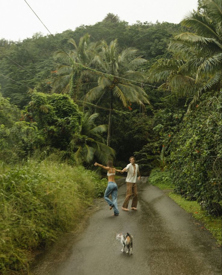 two people and a dog walking down a dirt road in front of some palm trees