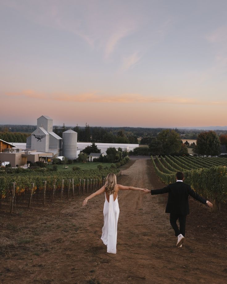 a man and woman holding hands walking down a dirt road in front of a vineyard