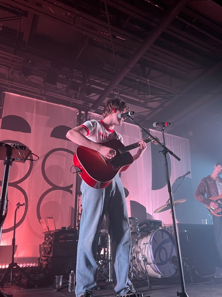 a man standing on top of a stage with a guitar in front of him and two microphones behind him