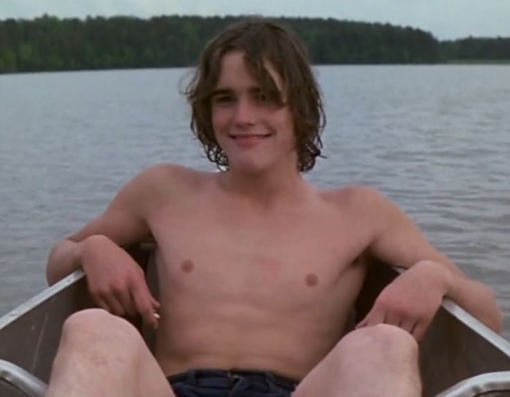 a shirtless young man is sitting in a boat on the water with his legs crossed