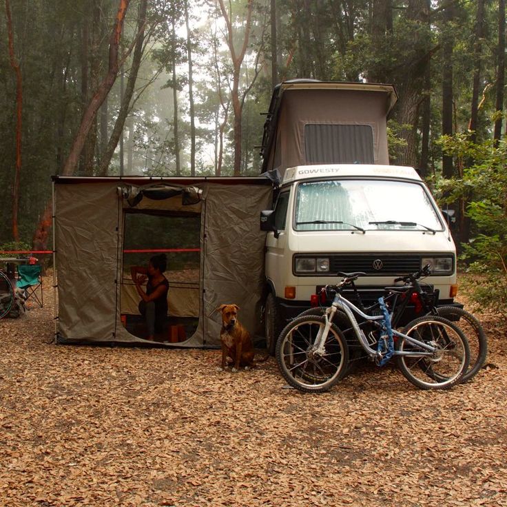 a van parked in the woods next to a bike and tent with a dog sitting outside