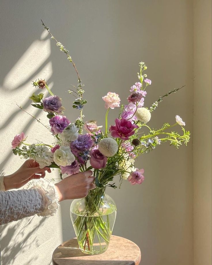 a woman arranging flowers in a vase on a small wooden table next to a white wall