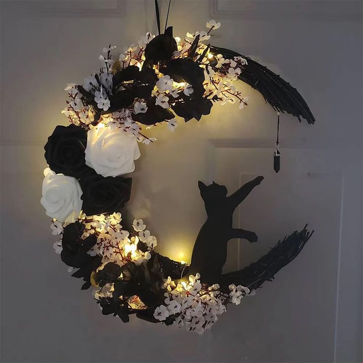 a black cat sitting on top of a wreath with flowers and lights in it's center