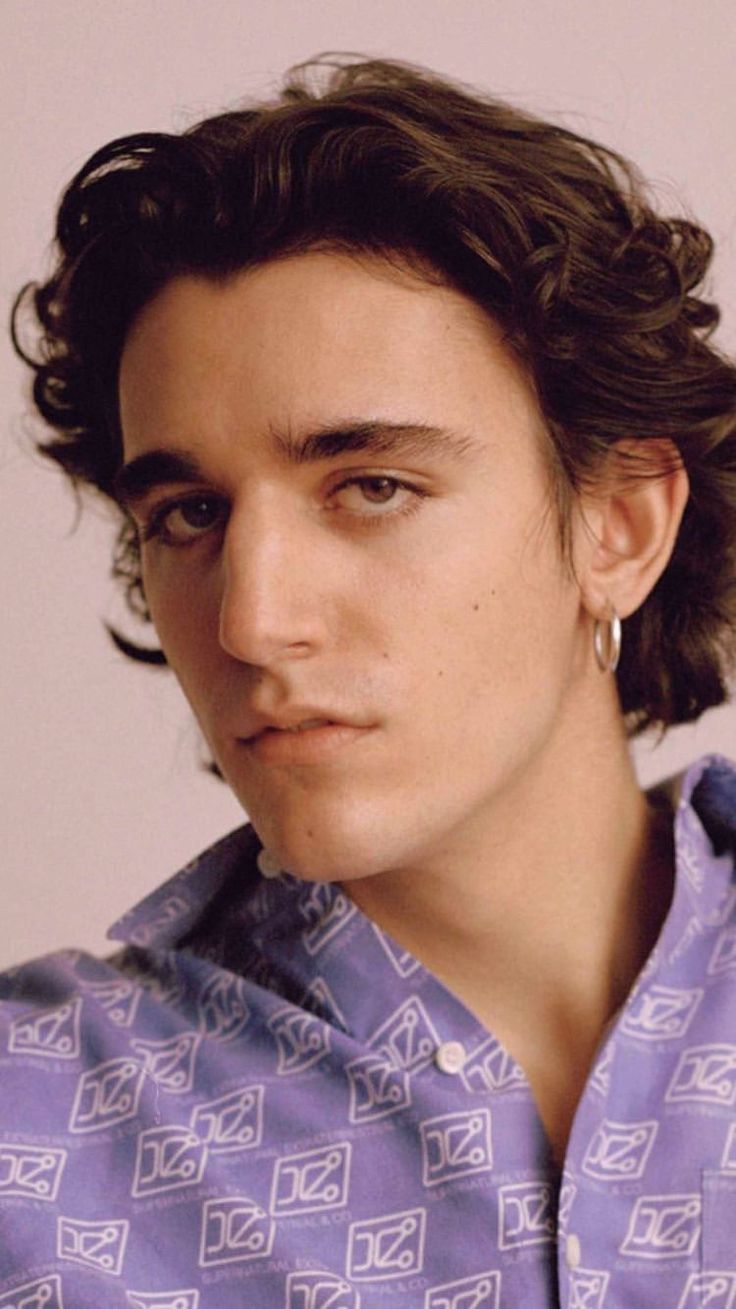 a young man with curly hair wearing a blue shirt and silver earrings looking at the camera