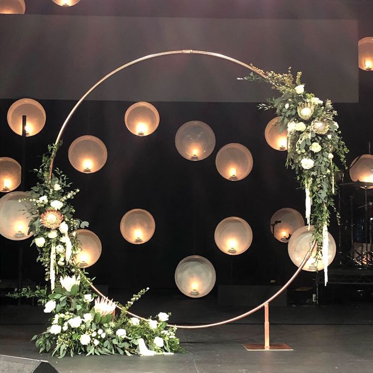 a circular arch decorated with flowers and greenery in front of a stage backdrop for a wedding ceremony