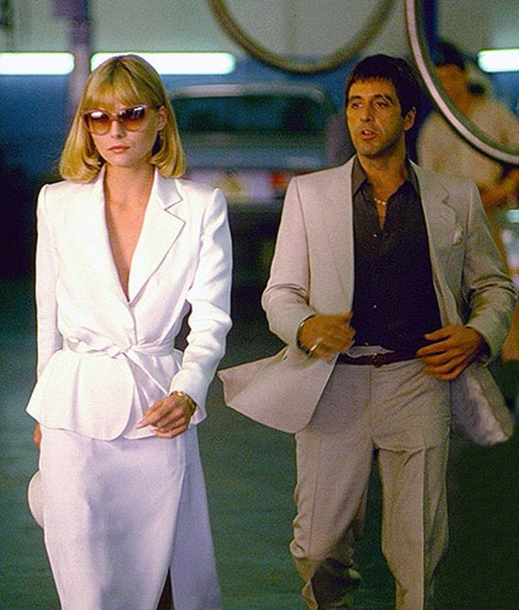 a man and woman are walking down the street in front of some cars, one is wearing a white suit