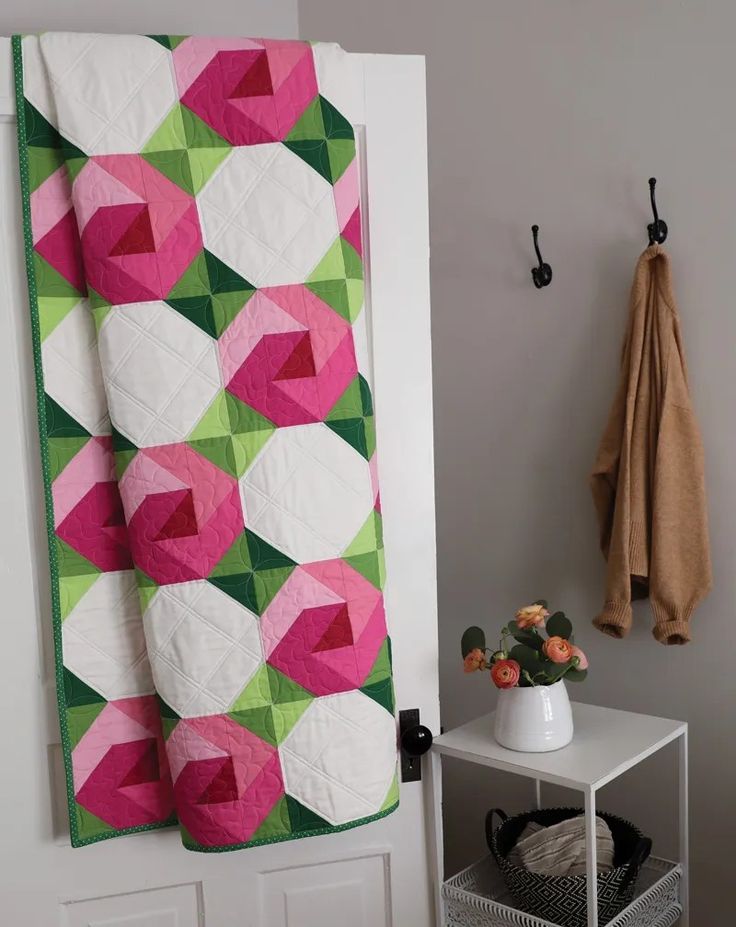 a pink and green quilt hanging on the wall next to a white table with flowers