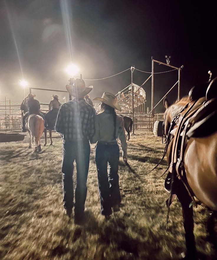 two men in cowboy hats are walking with horses on the field at night, while another man is standing next to them