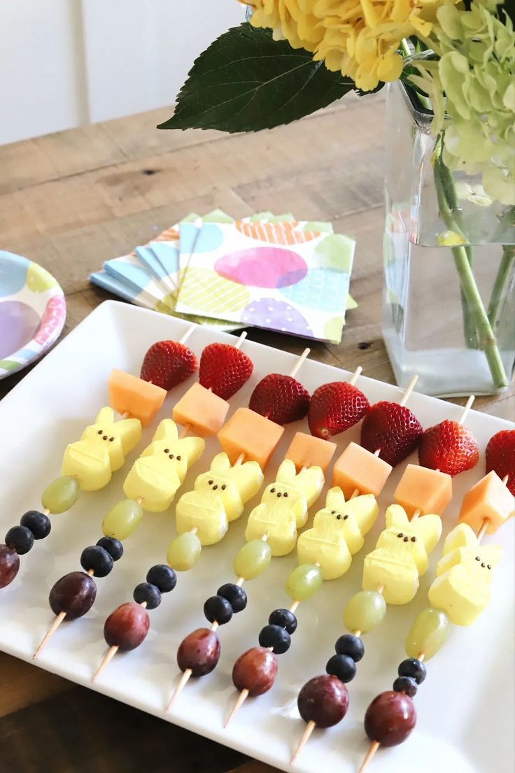 an assortment of fruit and cheese on skewers with flowers in vase behind them