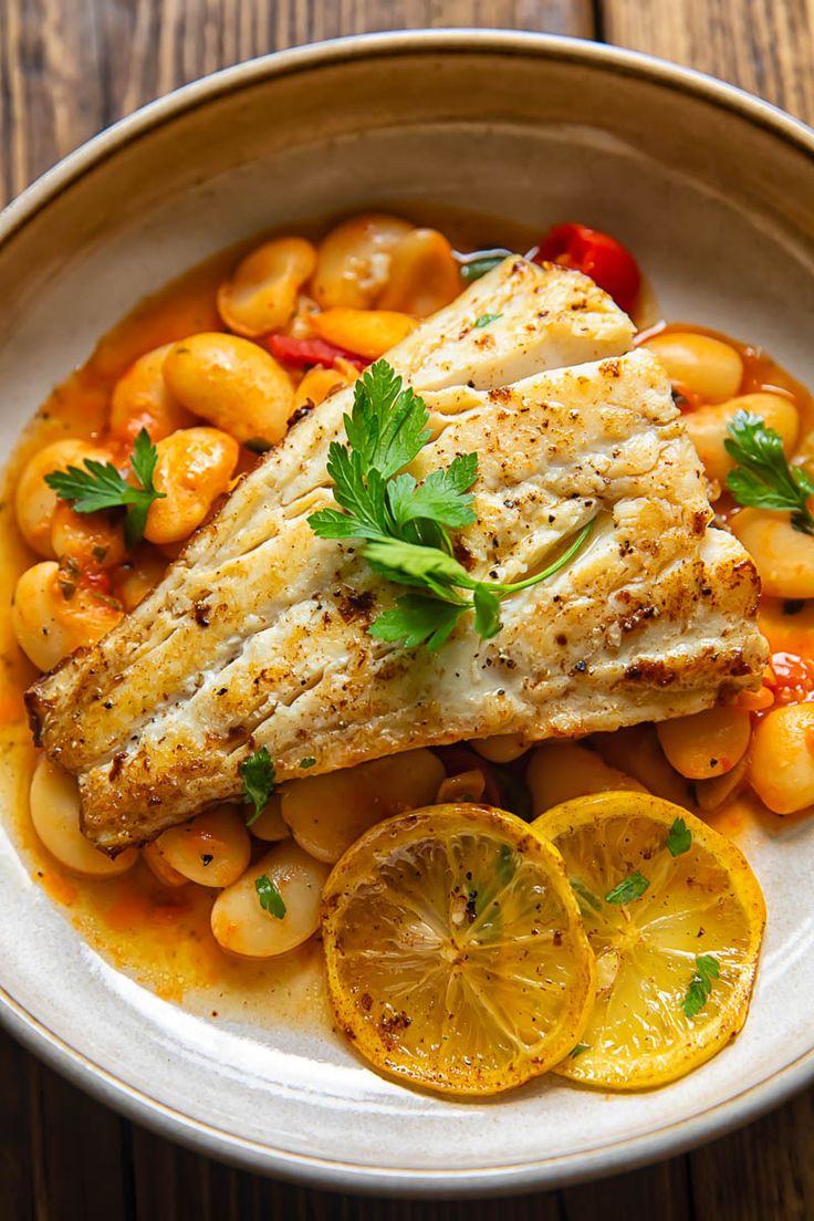 a white plate topped with fish, beans and lemons on top of a wooden table