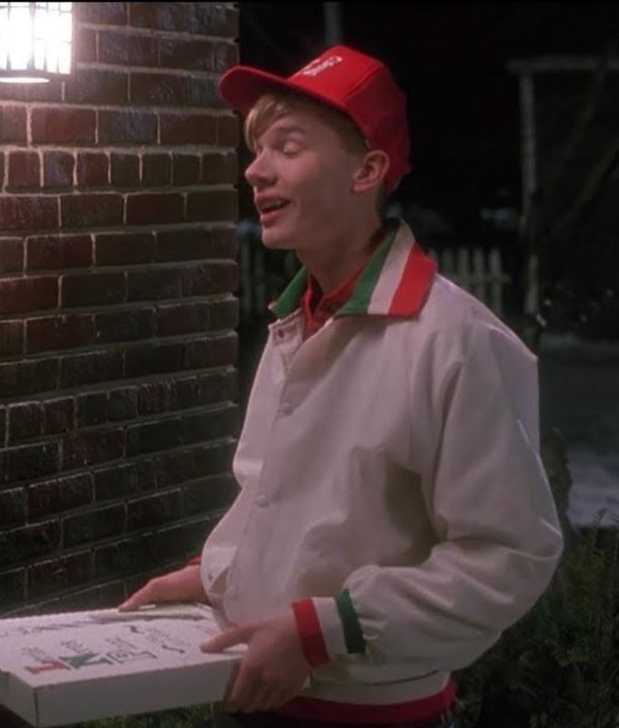 a man standing next to a brick wall wearing a red and white jacket with a green stripe around his neck