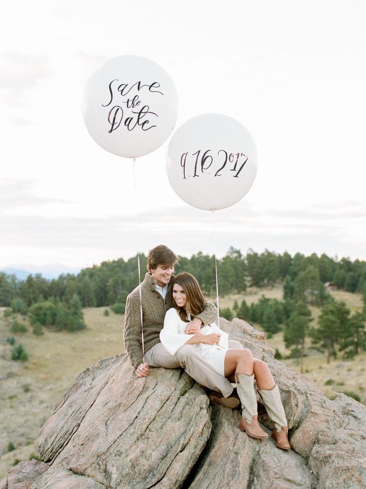 a man and woman sitting on top of a rock with two balloons in the air