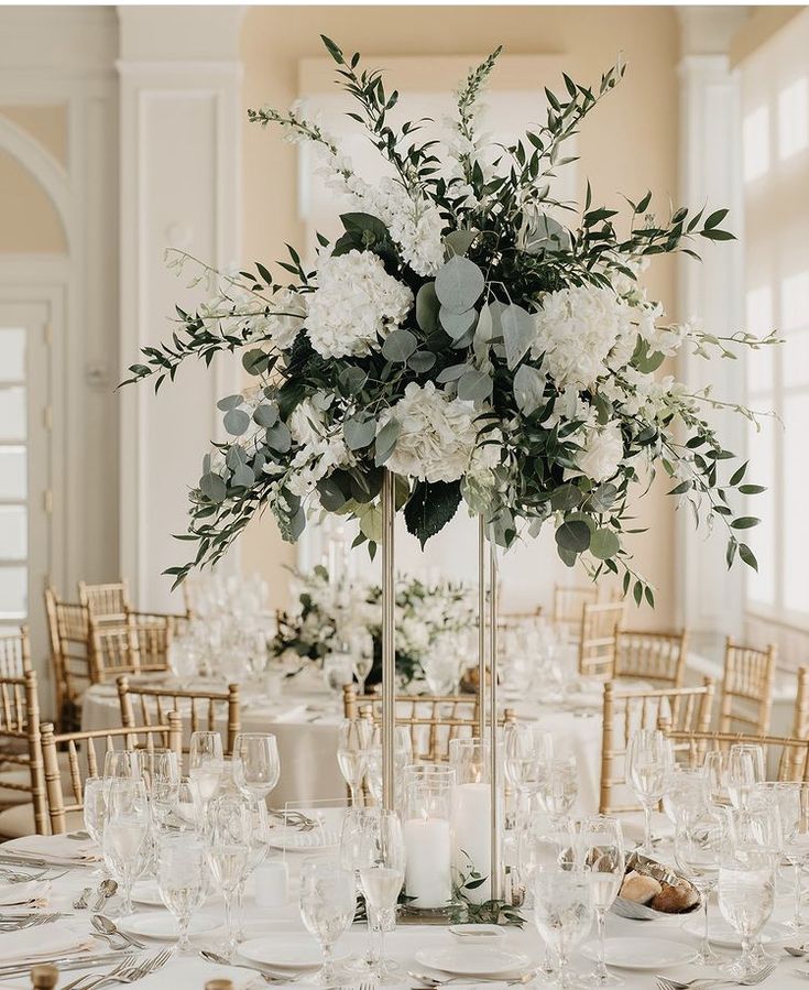 a tall vase filled with white flowers and greenery on top of a dining room table