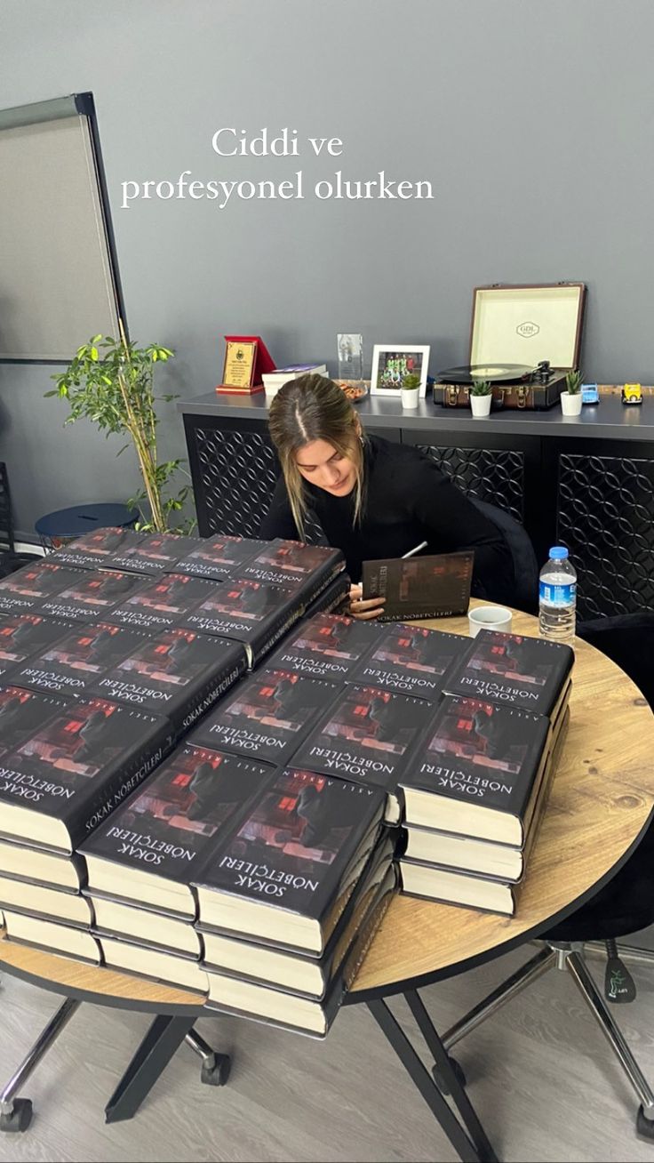 a woman sitting at a table with stacks of books on top of it and writing