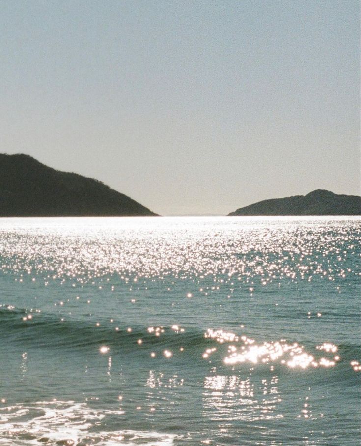 the sun shines on the water and hills in the distance, as seen from an ocean shore