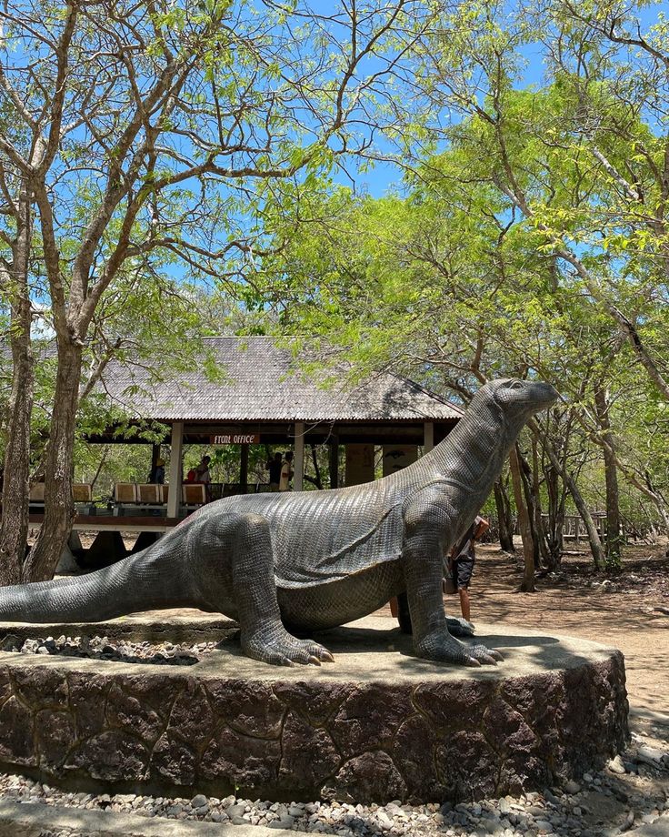 a statue of a dinosaur in the middle of a park with trees and people looking at it