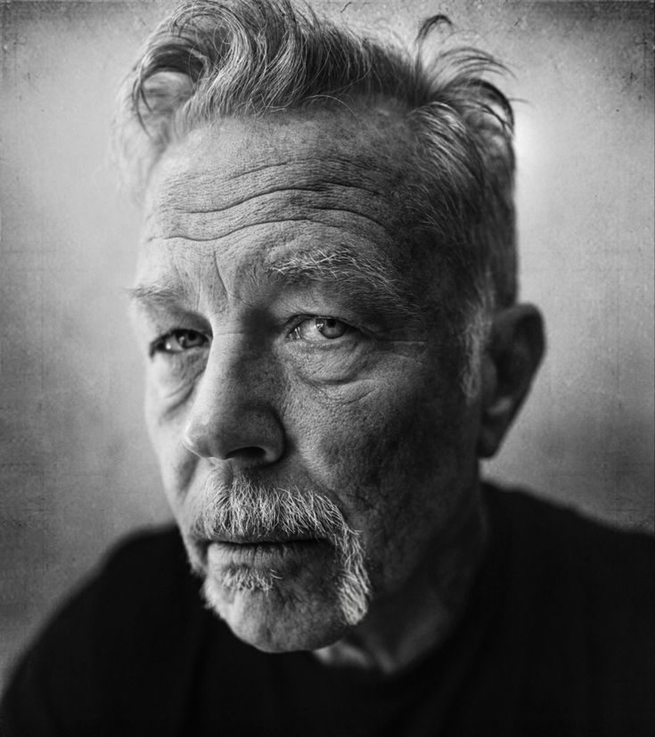 black and white photograph of an older man with grey hair looking at the camera while wearing a dark t - shirt