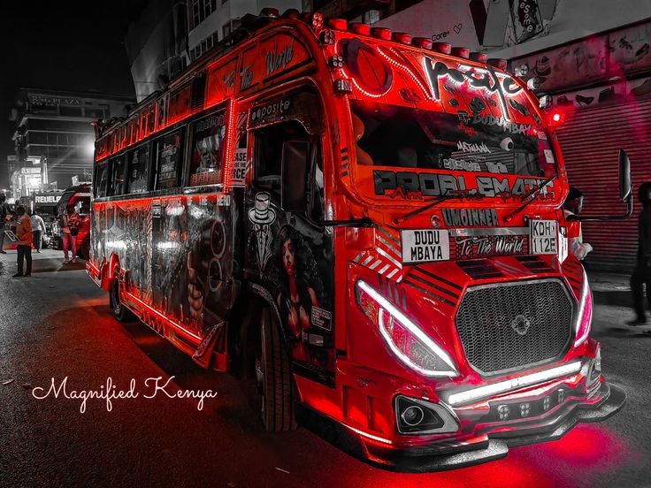 a red double decker bus parked in front of a building at night with people walking around