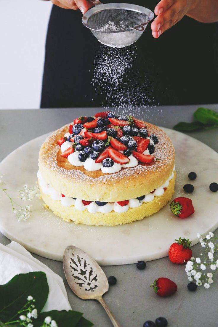 someone sprinkling sugar on top of a cake with berries and blueberries around it