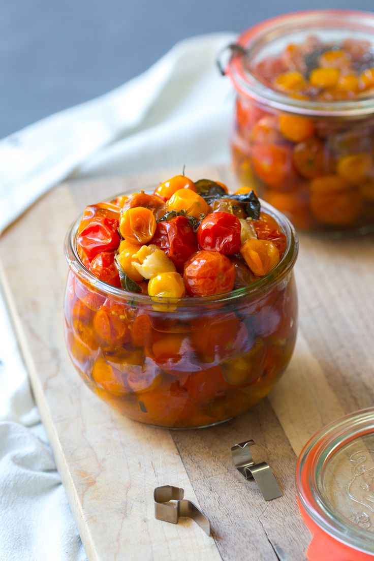 two glass jars filled with tomatoes and corn on top of a cutting board next to measuring spoons