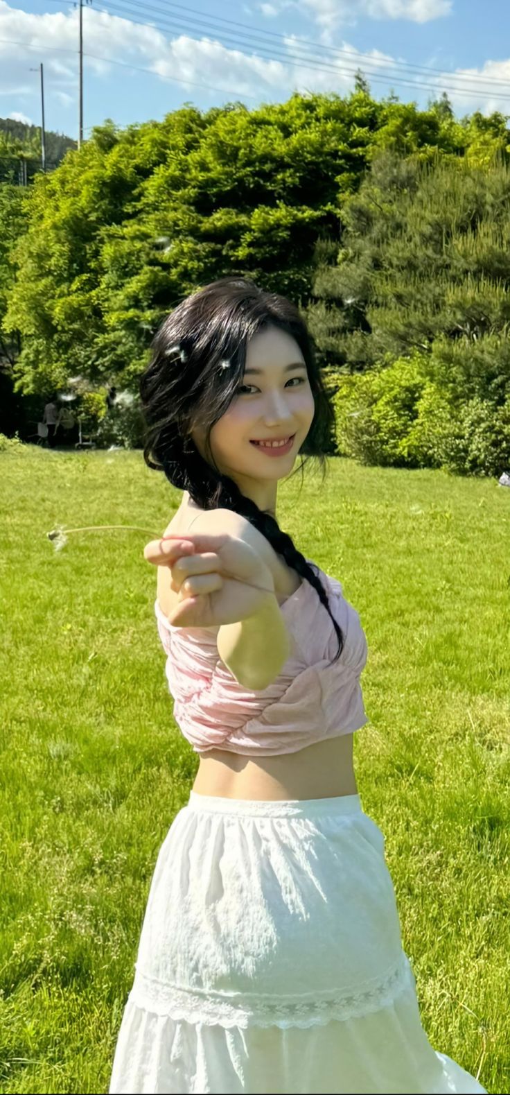 a woman in a white skirt pointing at the camera with trees in the back ground behind her