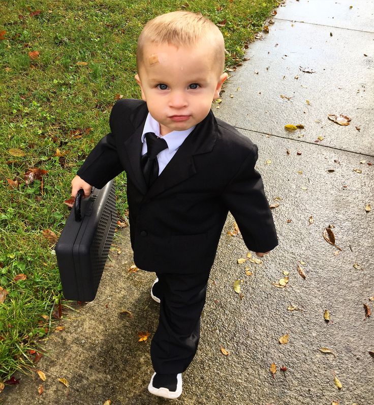 a little boy in a suit and tie walking down the sidewalk with a briefcase behind him