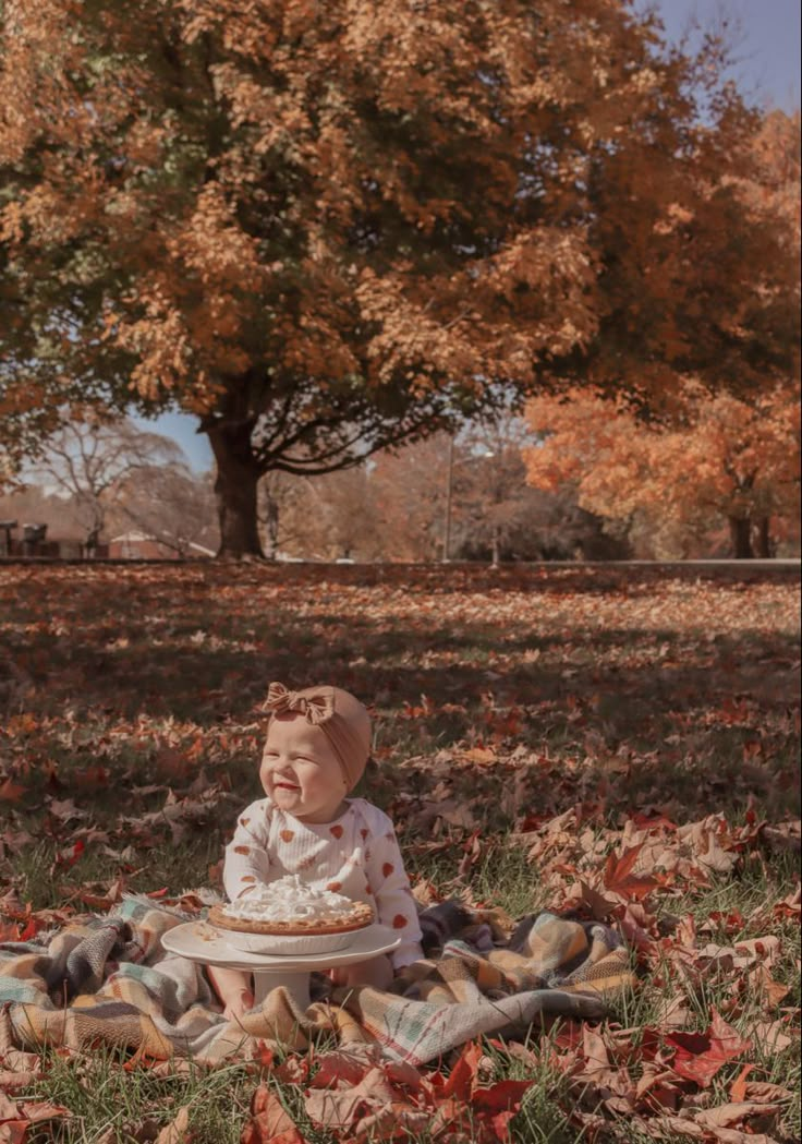 a baby sitting on top of a blanket in the grass next to a tree with leaves all over it