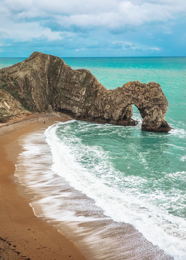 an arch shaped rock sticking out of the ocean next to a sandy beach with waves coming in