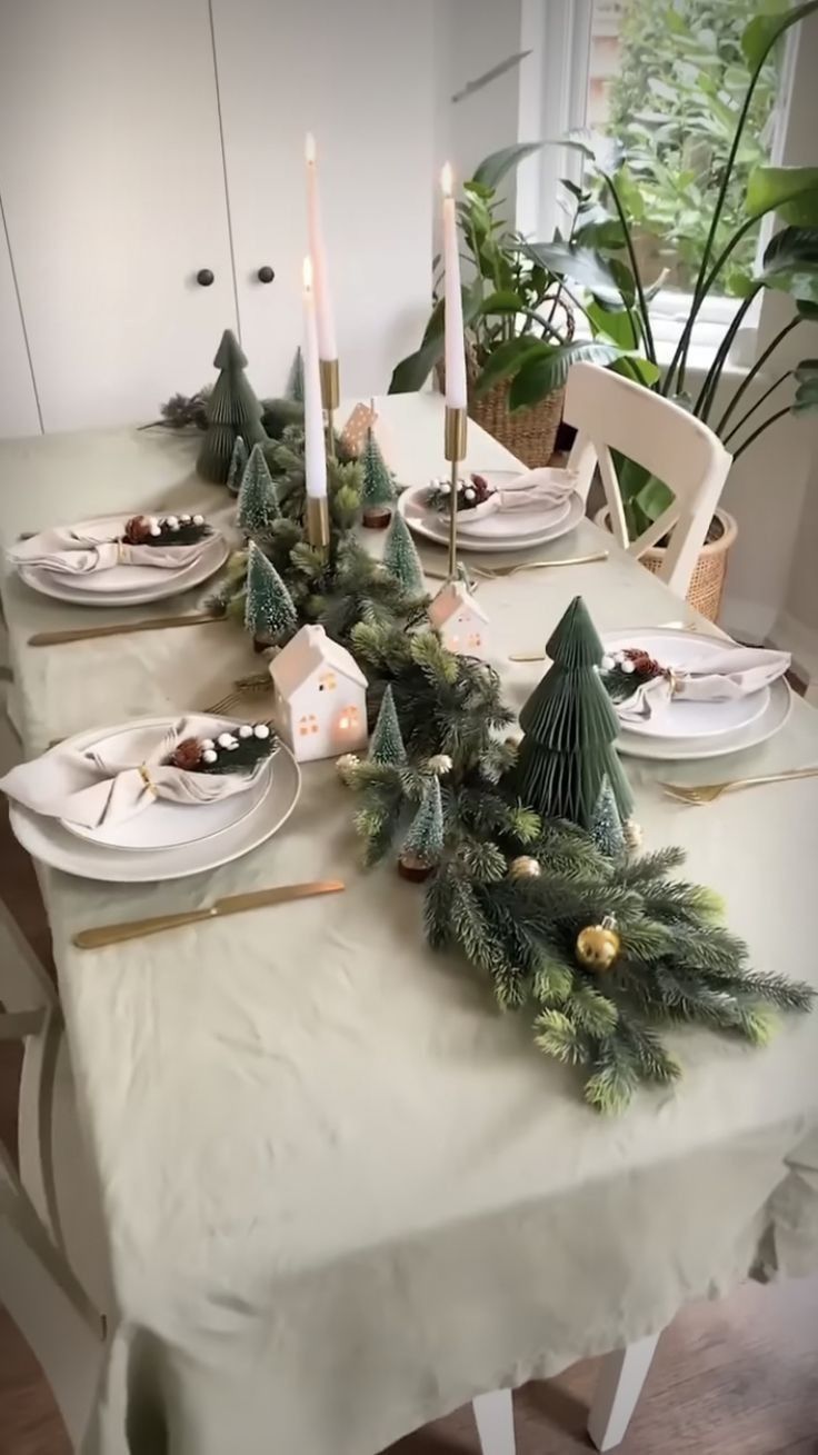 the table is set for christmas dinner with candles and greenery on it, along with other holiday decorations