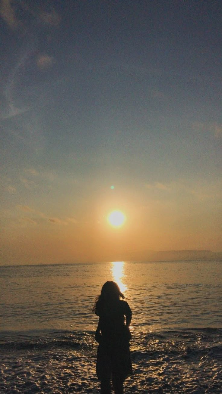 a woman standing on top of a sandy beach next to the ocean under a sunset