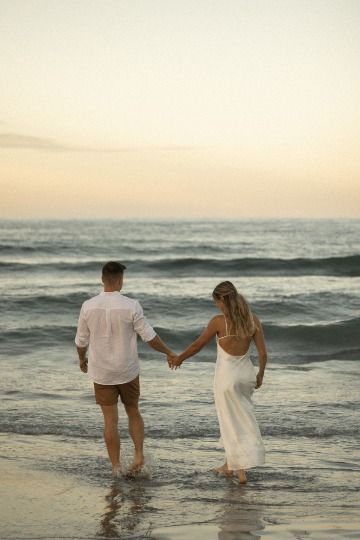 a man and woman holding hands while walking on the beach with waves in the background