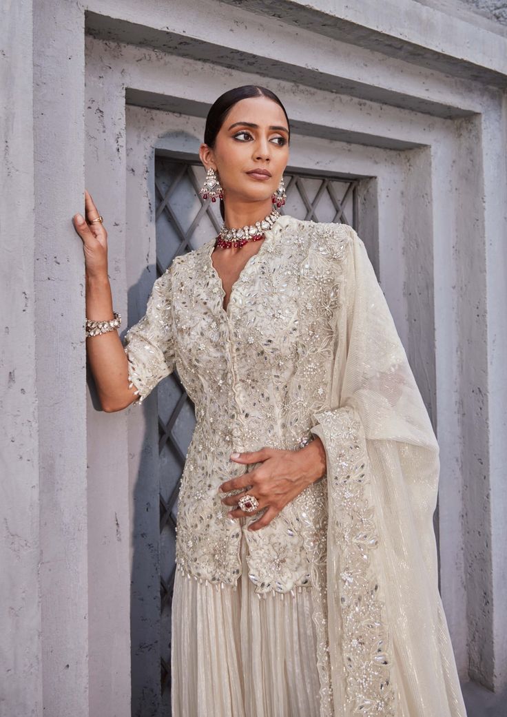 Step into elegance with this gorgeous ivory outfit featuring a machine-embroidered kurta with a hook-eye front opening. The crinkle fabric sharara adds a touch of sophistication to the ensemble. Completing the look is the dupatta, adorned with machine embroidery in borders. Embrace timeless beauty with this meticulously crafted outfit, designed to enhance your grace and style. White Chinon Sets For Eid, White Chinon Sets With Zari Work, White Sharara With Resham Embroidery In Chinon, White Chikankari Embroidered Sharara In Chinon, White Chinon Sharara With Resham Embroidery, White Anarkali Dress In Chinon, White Resham Embroidered Sharara In Chinon, White Sharara With Chikankari Embroidery, White Sharara With Intricate Embroidery For Eid